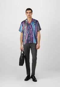 Short-sleeve shirt with a vibrant blue, pink, and black abstract pattern; gray trousers; black shoes; holding a black bag.