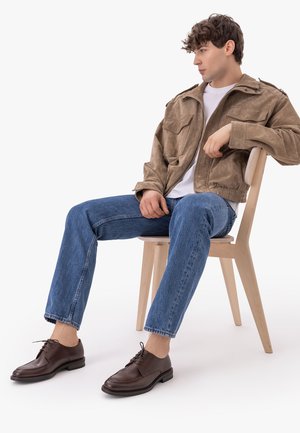 Young man sitting on wooden chair, wearing brown suede jacket, white shirt, blue jeans, and dark brown leather shoes.