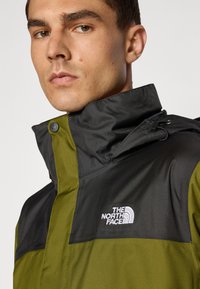 Herrejakke med en sort øverste sektion og olivengrøn nederste sektion, featuring en høj krave og The North Face-logoet i hvid.