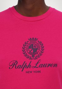 Lähikuva henkilöstä, joka käyttää kirkkaan vaaleanpunaista paitaa, jossa on musta Ralph Lauren -logo ja rintamerkki, jossa lukee "New York".