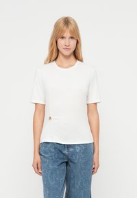 T-shirt blanc à manches courtes avec un col rond et un détail torsadé sur le côté, orné d'un bouton doré, associé à un jean en denim bleu à motif.