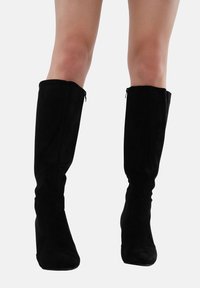 Botas negras de ante hasta la rodilla, con forma ajustada, punteras redondeadas y una cremallera lateral para facilitar su uso.