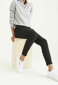 Felpa grigia con zip, pantaloni slim-fit neri e sneakers bianche. Posizione seduta su un blocco di legno chiaro, con uno sfondo neutro.
