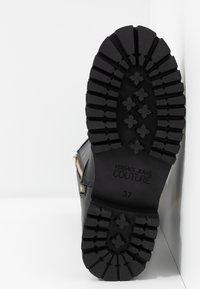 Czarna bota z teksturowaną gumową podeszwą z bieżnikiem, wytłoczonym logo "VERSACE JEANS COUTURE" oraz akcentem w postaci zamka błyskawicznego z boku.
