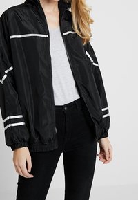 Chaqueta bomber negra con cremallera, franjas reflectantes, puños elásticos y un material ligero y brillante. Llevada sobre una camiseta gris.