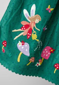Fee mit Flügeln und rotem Kleid, die bestickte, farbenfrohe Pilze und Blumen auf grünem Stoff mit kleinen blauen Punkten gießen.
