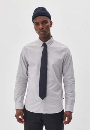 Grey button-up shirt with long sleeves, a pointed collar, and a navy tie. The fabric appears smooth with subtle texture. A navy knit beanie is worn.