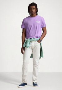 Μωβ βαμβακερό μπλουζάκι με λογότυπο "POLO", εκρού τζιν, πουλόβερ μέντας δεμένο γύρω από τη μέση και μπλε αθλητικά παπούτσια.