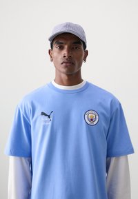 Šviesiai mėlynas džersis su apvalia iškirpte, siuvinėtu „Manchester City“ logotipu bei juodais „Puma“ ir „KidSuper Studios“ logotipais. Pora su dryžuotu kepurėle.