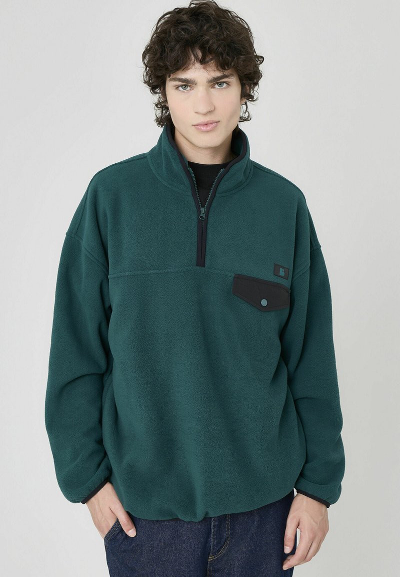 Pull-over polaire vert avec un col zippé mi-hauteur, une poche poitrine noire et des accents noirs contrastants. Texture douce et coupe décontractée.