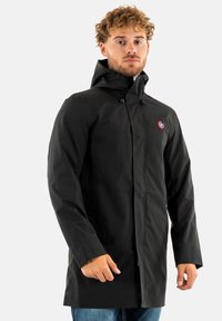 Manteau imperméable noir avec capuche, doté d'un zip à l'avant, de poches latérales et d'un petit logo rond sur la poitrine gauche.