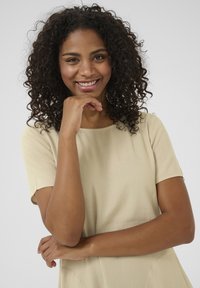 Top beige a maniche corte con scollatura rotonda, realizzato in tessuto morbido. Il modello sta sorridendo con la mano appoggiata sotto il mento, con capelli ricci.