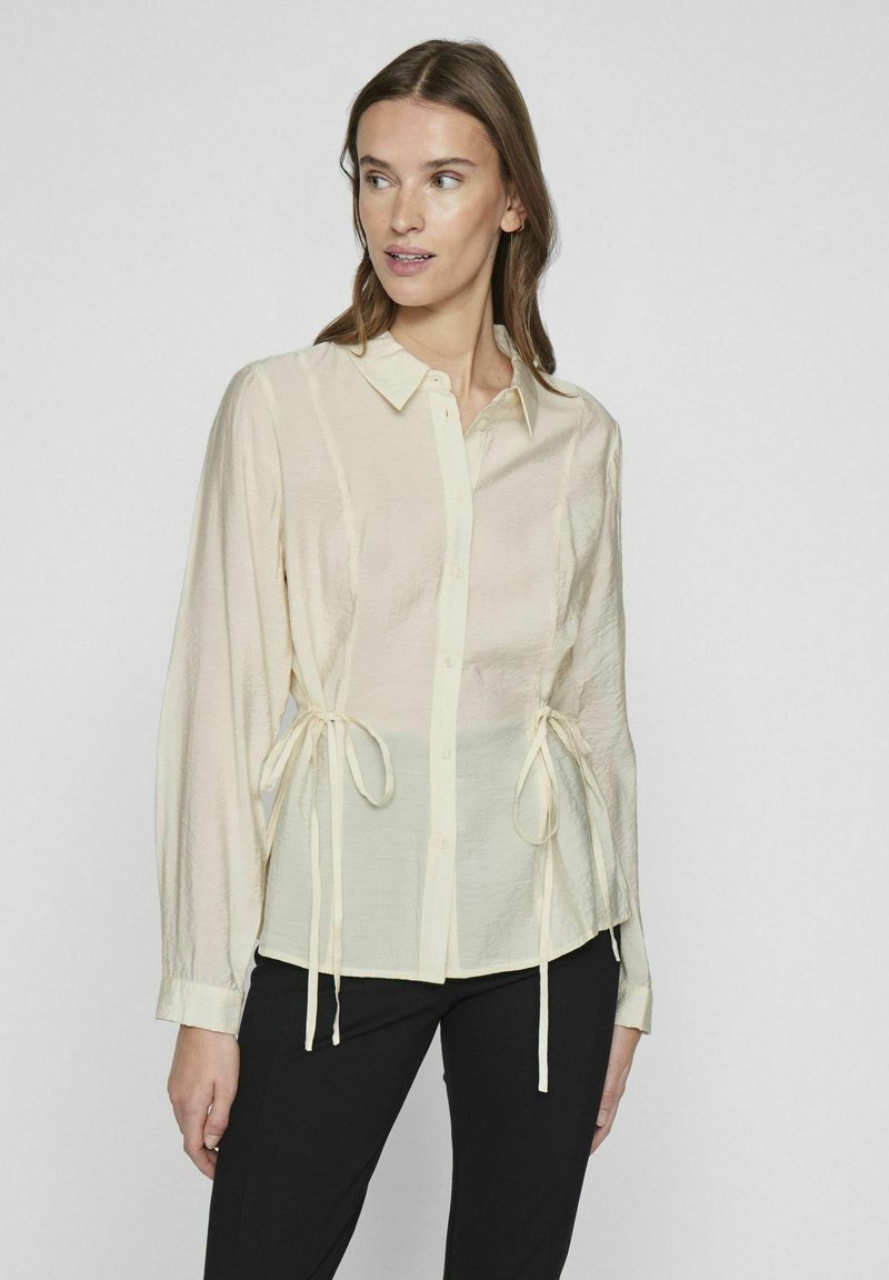 Chemise légère crème à manches longues avec une finition texturée, nouée à la taille avec des détails en ruban. Design de col classique.