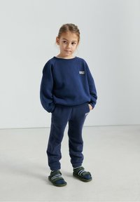 Laivastonsininen fleece-collegepaita pyöreällä kaula-aukolla ja logolla. Samaa laivastonsinistä rentoävyiset collegehousut. Yhdistetty tummiin sandaaleihin ja sukkiin.