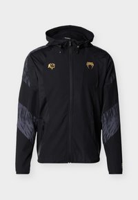 Μαύρο αδιάβροχο μπουφάν με κουκούλα, με χρυσά λογότυπα, γκρι διακοσμητικές λεπτομέρειες, φερμουάρ σε τσέπες και μοντέρνο αθλητικό σχέδιο.