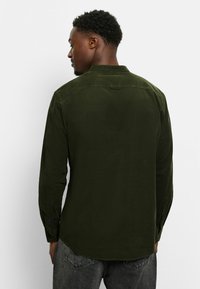 Langärmliges, dunkelolivgrünes Hemd aus strukturiertem Stoff, mit Kragen, geknöpften Manschetten und abgerundetem Saum. Verfügt über minimalistische Details.