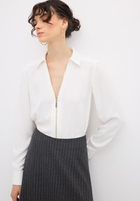 Blusa bianca con zip, maniche lunghe e colletto, abbinata a una gonna a righe verticali grigio scuro. Texture del tessuto morbida e dettagli di design eleganti.