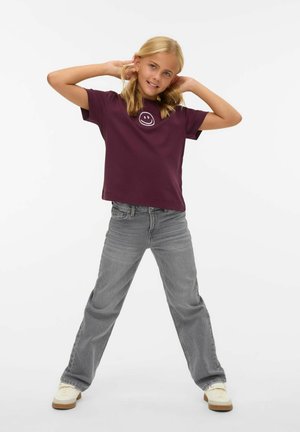 Ung pige står med benene fra hinanden, hænderne bag hovedet, iført en lilla t-shirt med et smilende ansigt og grå jeans mod en hvid baggrund.