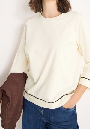 Frau trägt cremefarbenes Langarmshirt mit schwarzem Besatz und blaue Jeans, hält eine gefaltete braune Jacke in einer Hand, andere Hand in der Hosentasche.