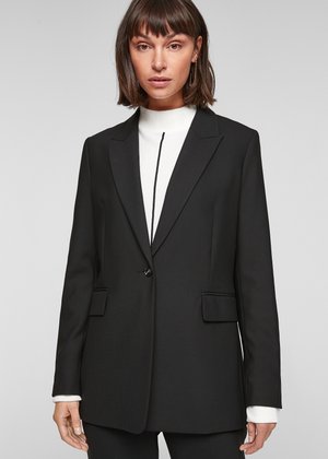 Blazer noir ajusté avec un design structuré, fermeture à un seul bouton et deux poches avant. Porté sur un haut blanc à col montant.
