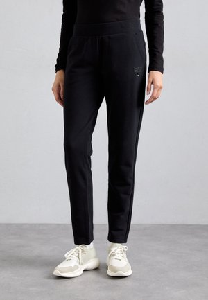 Personne portant un pantalon de survêtement noir de marque EA7 et des baskets blanches, debout sur un sol gris contre un mur gris clair.