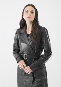 Žena s dlouhými tmavými vlasy, oblečená v černém koženém trenčkotě s páskem, stojící před jednoduchým bílým pozadím.