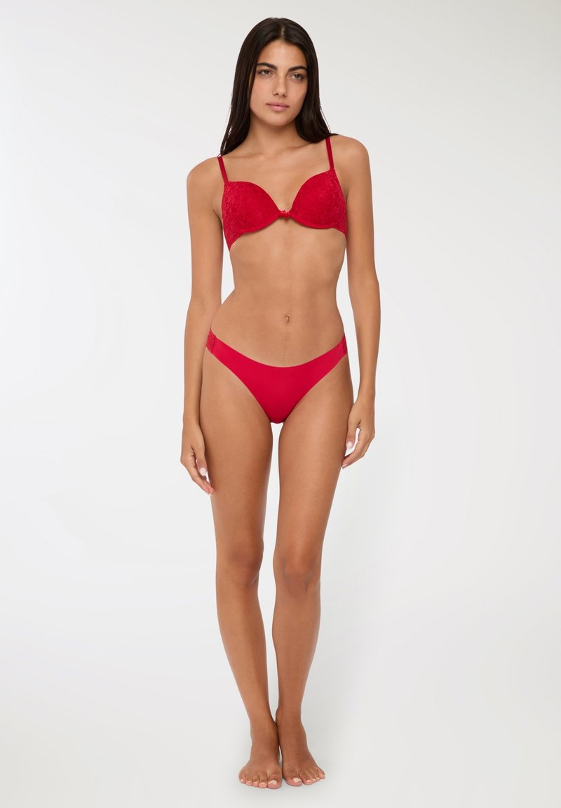 Reggiseno push-up in pizzo rosso con slip coordinati. Il reggiseno presenta una finitura liscia con un fiocco centrale e spalline regolabili.