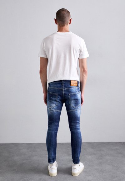 Man wearing white t-shirt, slim-fit blue jeans with back pockets and leather patch, and white sneakers standing against plain gray wall.