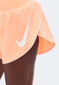 Αθλητής που φοράει ανοιχτό πορτοκαλί σορτς Nike για τρέξιμο με λευκή επένδυση, κοντινή λήψη που δείχνει το λογότυπο Nike swoosh σε μηρό με σκουρόχρωμο δέρμα.