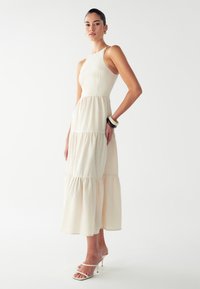 Nainen ilman hihoja kerroksittain vaaleassa kermakuisessa midi-mekossa, joka on pukeutunut hihnallisiin korkokenkiin ja paksuihin koruihin valkoista taustaa vasten.