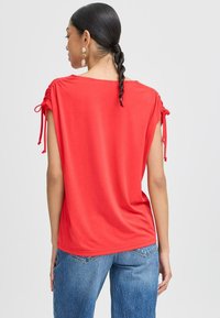 Rode top met korte mouwen, met geplooide schouderdetails, vloeiende silhouette en zachte textuur. Draagt een lichtblauwe high-waisted jeans.
