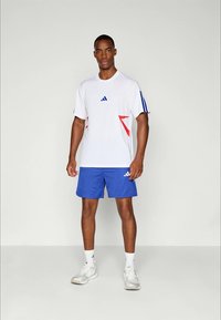 Man in een wit sporthemd met blauwe en rode accenten, blauwe shorts, witte sokken en witte sneakers, staande tegen een effen lichte achtergrond.