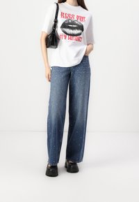 T-shirt grafico bianco con labbra nere in evidenza e testo rosso. Jeans in denim a gamba larga blu adornati con borchie argentate. Scarpe chunky nere.