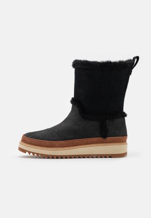 Czarny zimowy but sięgający do połowy łydki z futrzanym obszyciem, brązowymi zamszowymi akcentami, beżową grubą podeszwą i karbowaną podeszwą zewnętrzną zapewniającą przyczepność.