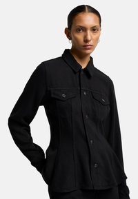 Camicia nera con bottoni, realizzata in tessuto resistente, caratterizzata da un design aderente, colletto appuntito e tasche frontali con chiusura a bottone.