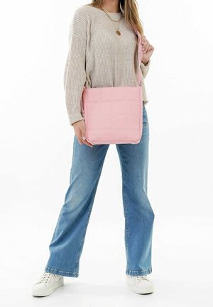 Frau, die einen beigen Pullover, blaue Jeans und weiße Sneakers trägt, während sie eine rosafarbene Schultertasche mit gestepptem Muster über den Körper trägt.