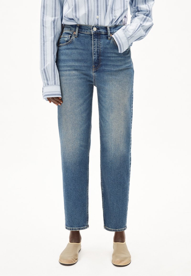 Personne portant un jean bleu taille haute et une chemise à rayures bleu clair à manches longues, une main dans la poche et des chaussures beiges sans lacets.