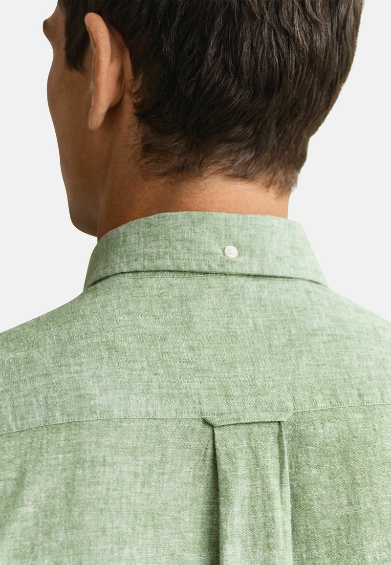 Mand set bagfra iført en lys grøn hørskjorte med knaplukket krave og detalje med bagplissering mod en ensfarvet baggrund.