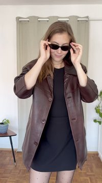 Καφέ δερμάτινο oversized μπουφάν με μεγάλους κομψούς, μαύρη φόρμα και μαύρα γυαλιά ηλίου. Υφή υφάσματος και χαλαρή εφαρμογή, σε ανοιχτό φόντο.