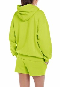 Felpa strutturata verde neon con un grande cappuccio, abbinata a pantaloni corti coordinati. Entrambi i capi presentano un design casual e morbido.