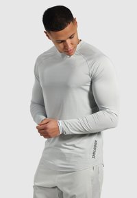 Helles graues langärmliges Sportshirt mit einer figurbetonten Passform, glatter Textur und kontrastierendem Logo an der Seite. Flache Nähte für mehr Komfort.