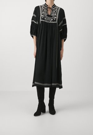 Kvinde iført en løs sort midi-kjole med hvid blomsterbroderi, trekvartærmer og sorte støvler med hæl, stående foran en ensfarvet baggrund.