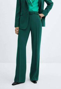 Groen op maat gemaakt pak met een getailleerde blazer en wijde broeken. De stof heeft een gladde textuur, met een bijpassende ceintuur en gepolijste zwarte schoenen.