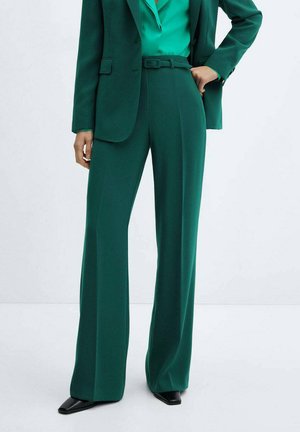 Groen op maat gemaakt pak met een getailleerde blazer en wijde broeken. De stof heeft een gladde textuur, met een bijpassende ceintuur en gepolijste zwarte schoenen.