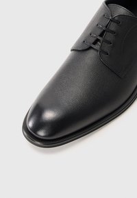 Zapato de vestir de cuero negro pulido con superficie texturizada y cordones cerrados, fotografiado sobre un fondo gris claro.