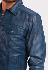 Modrá kožená bunda s výrazným límcem, předním zipem, dvěma náprsními kapsami se zapínáním na patentky a hladkou texturou s jemným leskem.