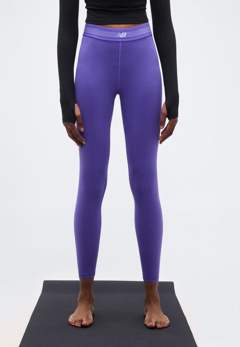 Άτομο που φοράει μοβ κολάν New Balance και μαύρο μακρυμάνικο μπλουζάκι, όρθιο ξυπόλυτο σε μαύρο στρώμα γιόγκα.