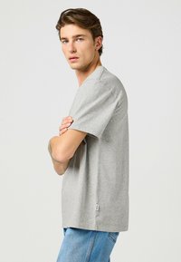 Camiseta gris de cuello redondo hecha de tejido suave, con mangas cortas y un corte relajado. Llevada con vaqueros azules, mostrando una vista lateral.