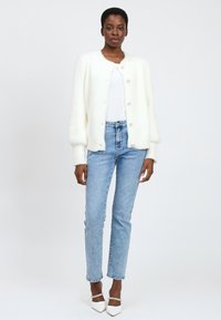 Vrouw in een witte harige knopencardigan, witte top, lichtblauwe spijkerbroek en witte puntige hakken met enkelbandjes, staande tegen een effen achtergrond.
