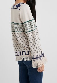 Pull en tricot présentant des motifs géométriques en beige, vert et bleu marine, avec un col rond, des manches évasées et un ourlet à franges.